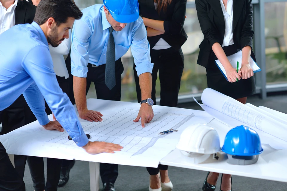 Réunion de travail en intérieur avec équipe de professionnels analysant des plans architecturaux sur table blanche